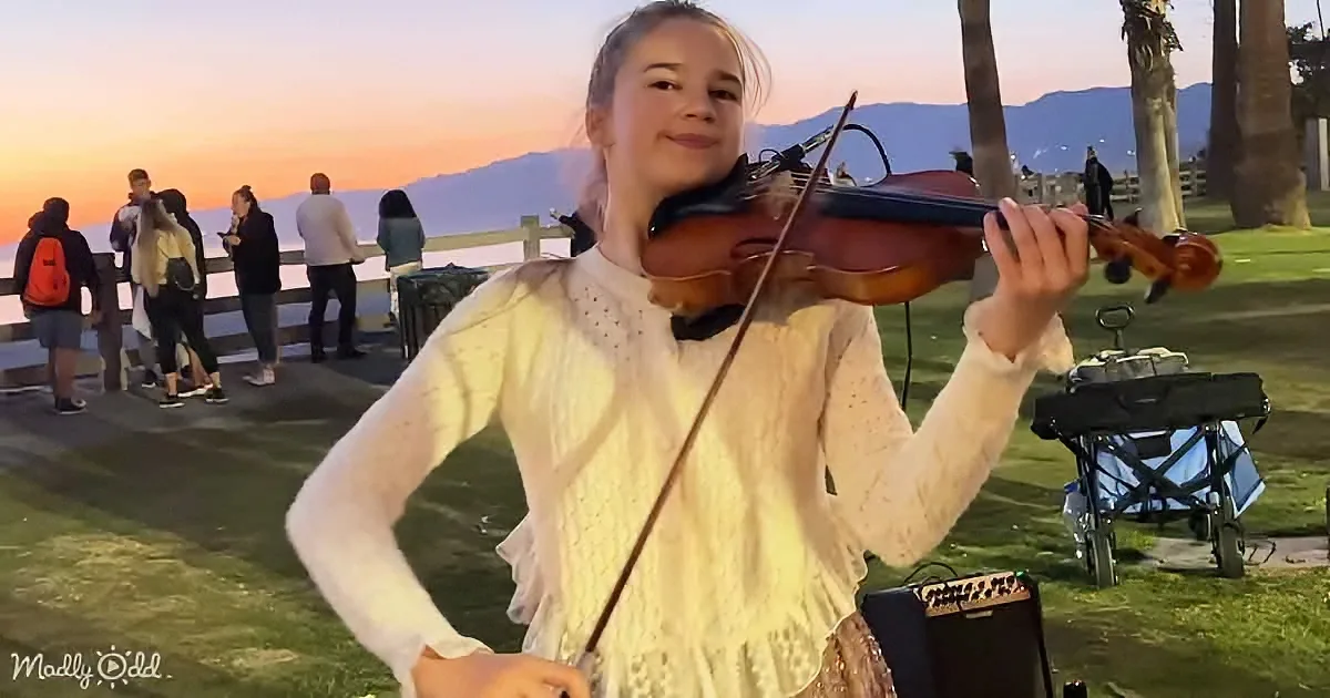 A girl in a white dress plays the violin at sunset near a grassy area with people in the background.