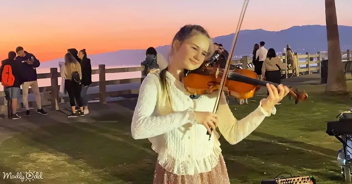 A girl in white plays violin at sunset, surrounded by people on a grassy area near a railing.