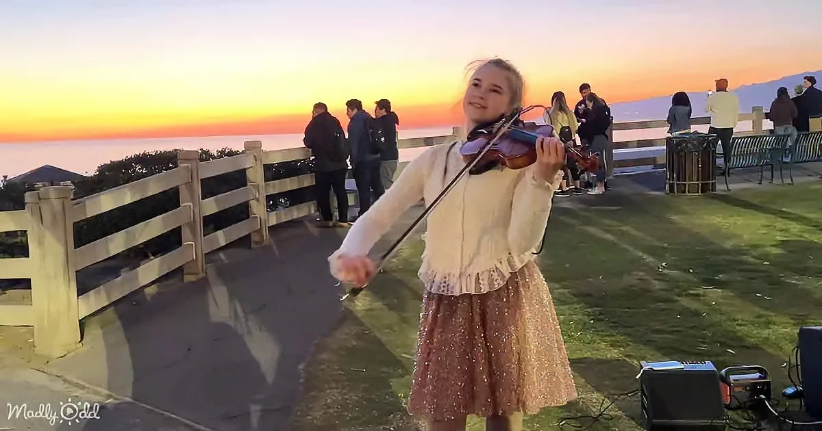 A young woman plays violin at sunset, surrounded by people on a scenic overlook.