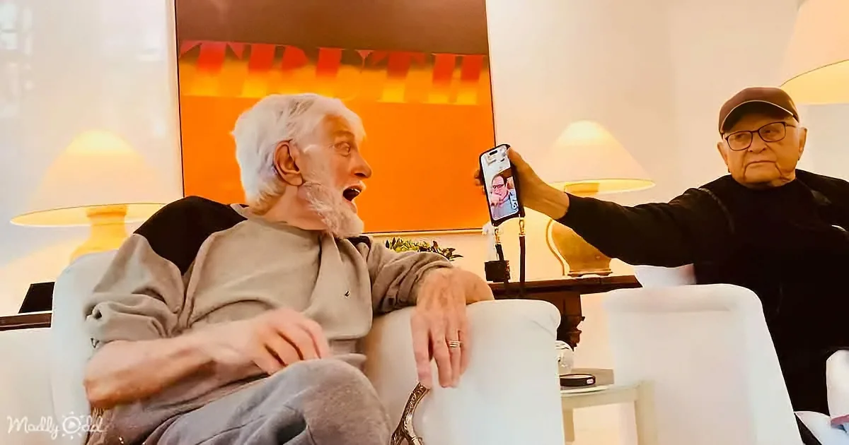 An elderly man sits on a couch, surprised as another holds up a phone showing a video call.