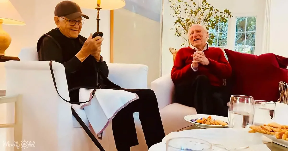 An older man in a black outfit sits on a white chair, holding a phone, while another man in a red sweater sits on a red...