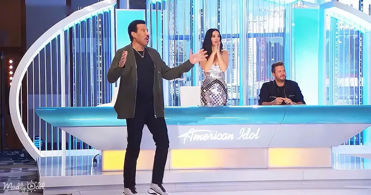 A man stands on stage at "American Idol," gesturing with his hands, while judges sit behind him.
