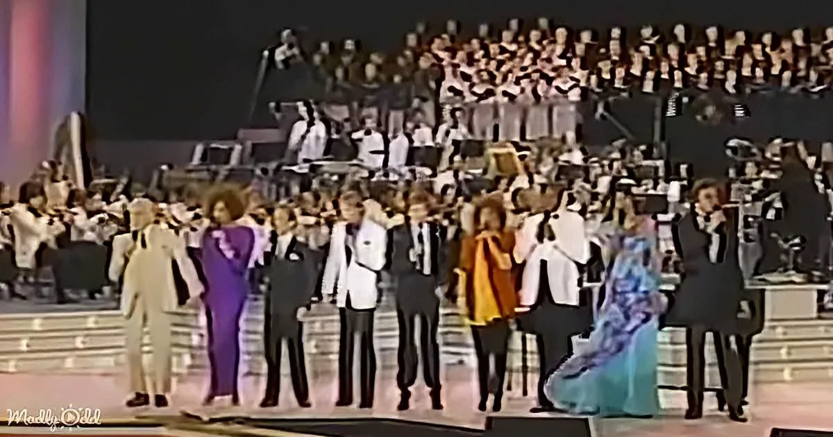A group of performers in formal attire stands on stage, facing a large choir and orchestra in the background.