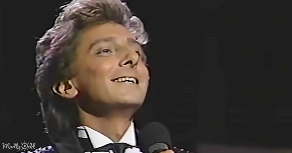 A person with short, curly hair and a white shirt smiles while holding a microphone.