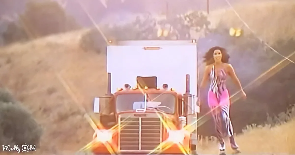 A woman in a pink dress walks confidently beside a large orange truck on a dusty road.