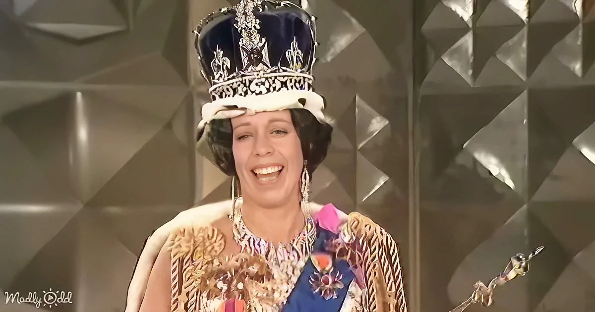 A person in a colorful, ornate outfit and crown smiles on stage, holding a trophy.
