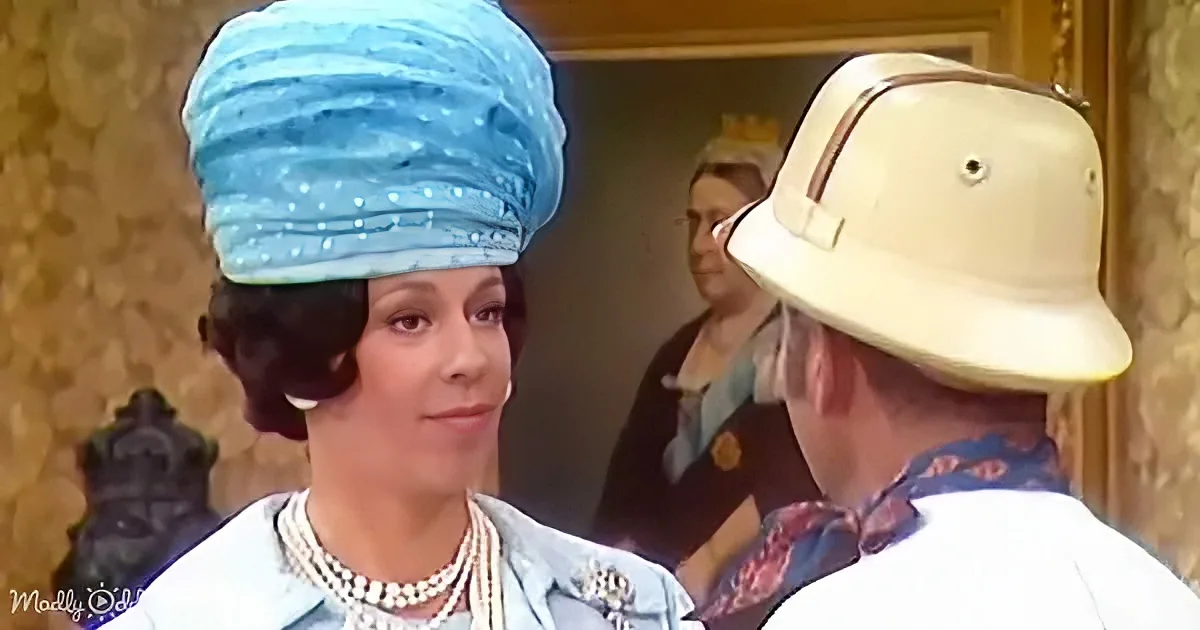 A woman in a blue turban and pearls smiles at a man in a white hat and scarf, with another woman in the background.
