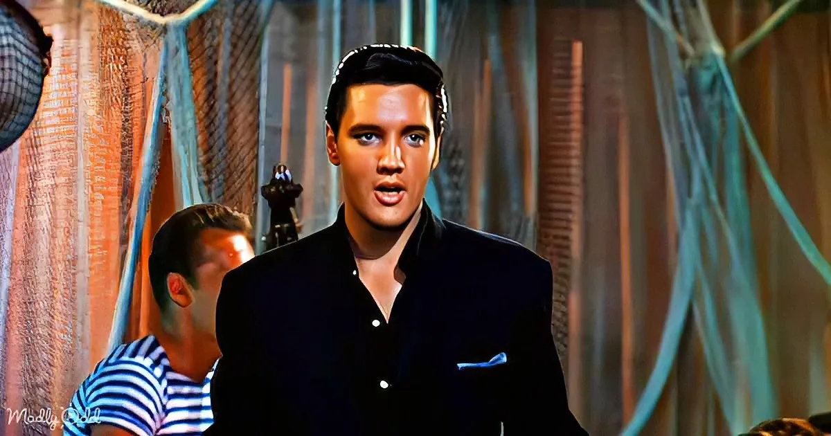 Elvis Presley in a black suit, mid-song, with a backdrop of draped fabric and a man in a striped shirt.