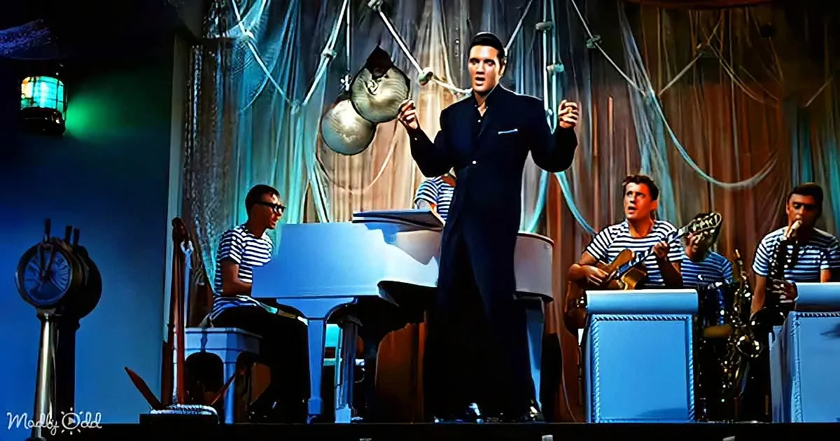 Elvis Presley in a black suit sings on stage with a band and a blue piano.
