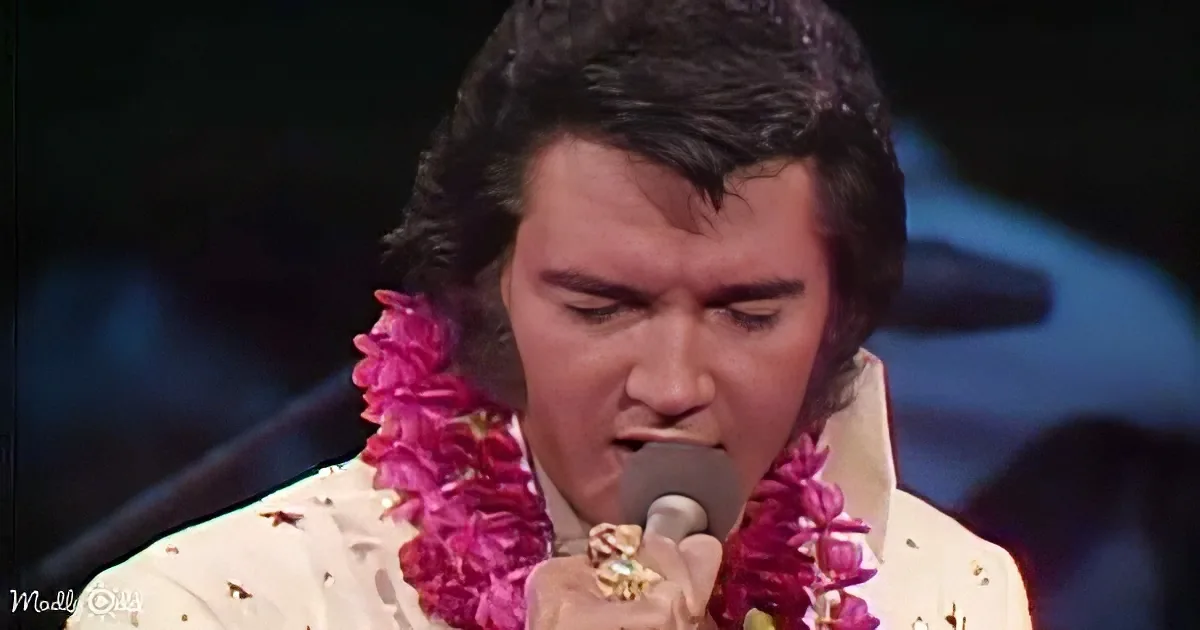 Elvis Presley singing into a microphone, adorned with a pink lei, on stage.
