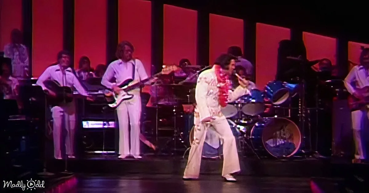 A performer in a white outfit with a red scarf is dancing on stage with a band in the background.