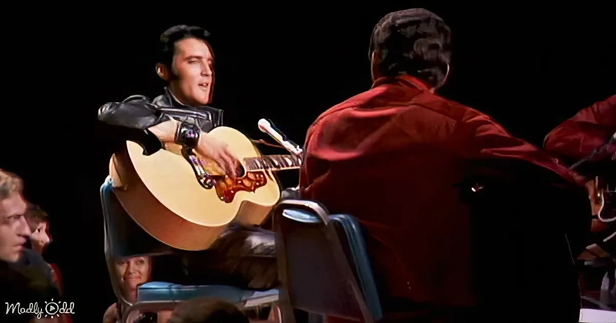 Elvis Presley performs on stage, holding a guitar, with a red-suited band member in the foreground.