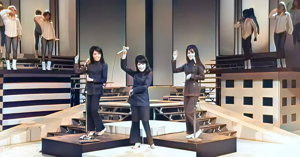Three women in black outfits perform on stage with microphones, surrounded by a building backdrop.