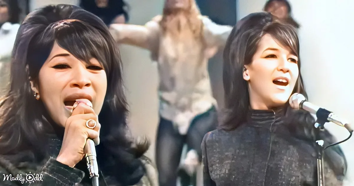 Two women sing into microphones, one with a ring, both wearing black, with a blurred crowd in the background.