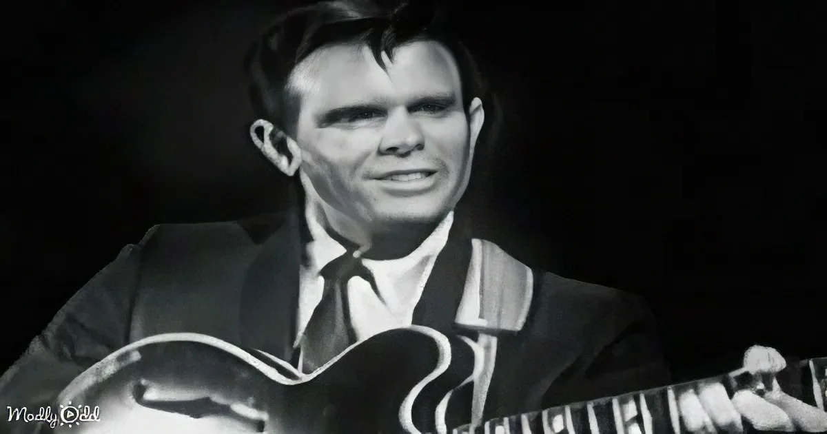 A man in a suit holds a guitar, smiling, in a black-and-white image.