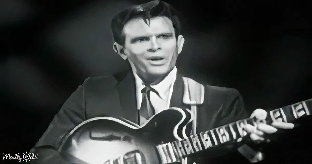 A man in a suit plays a guitar with a dark background.