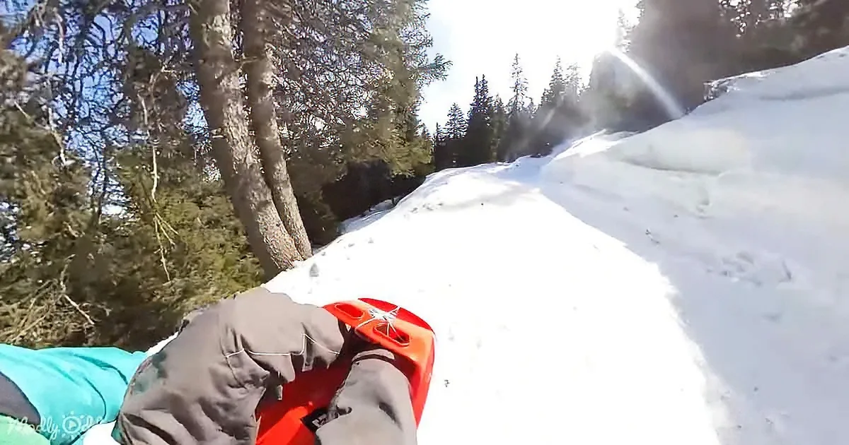 A person in a teal shirt and brown pants is sledding down a snowy hill with a red sled.
