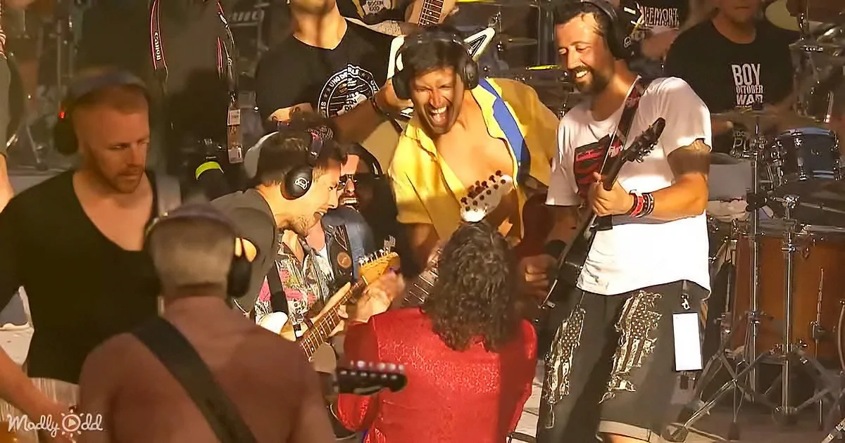 A man in a yellow shirt is passionately singing and playing guitar on stage with a band.