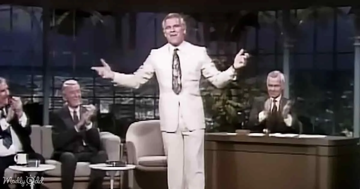 A man in a white suit stands with arms outstretched, smiling, while seated men clap in the background.