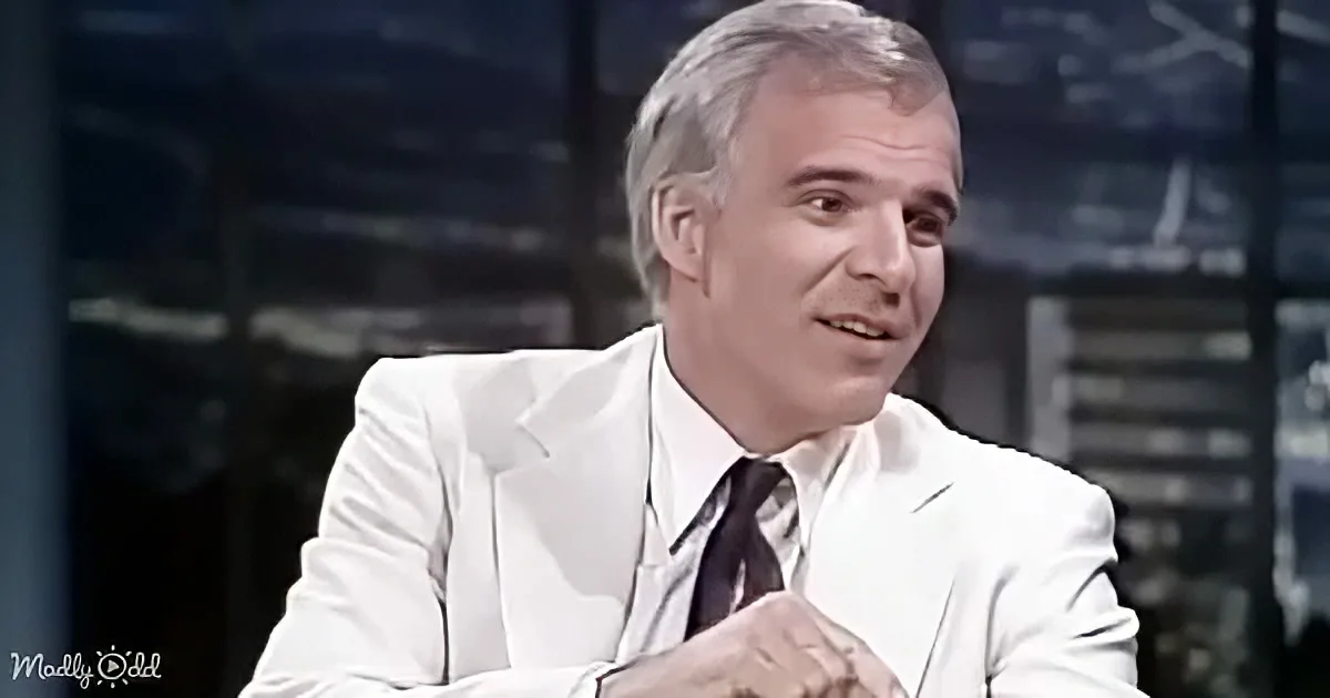 A man in a white suit and tie sits with hands clasped, looking to the side, in a softly lit room.