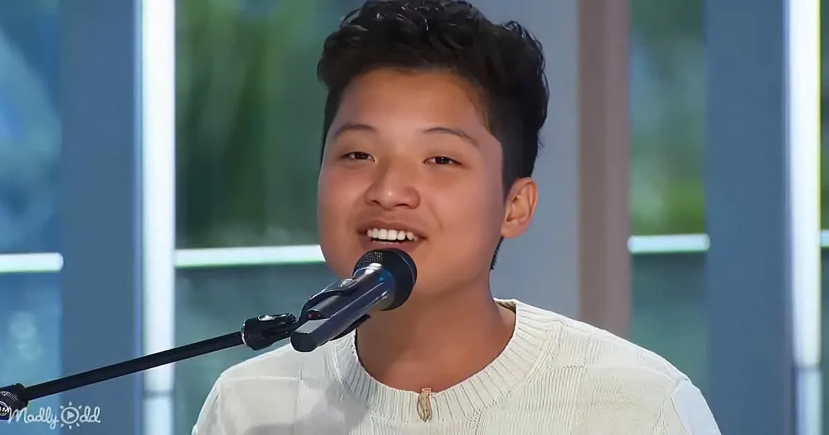 A young person smiles while singing into a microphone, wearing a white sweater.