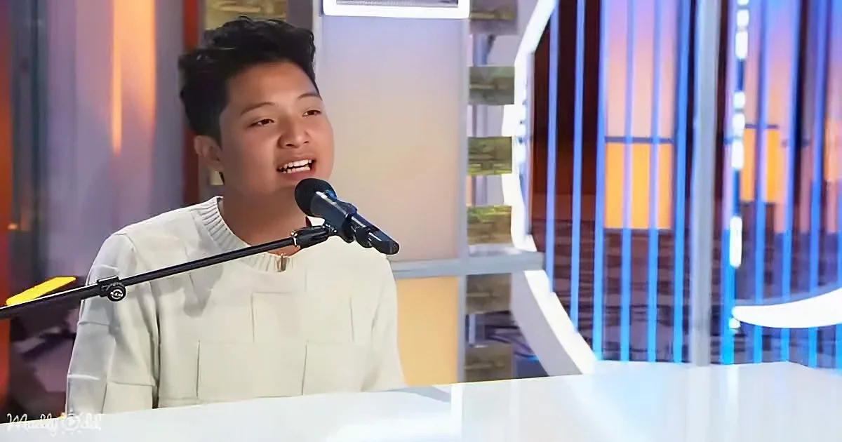 A young person in a white sweater sings into a microphone on a stage with colorful lighting.