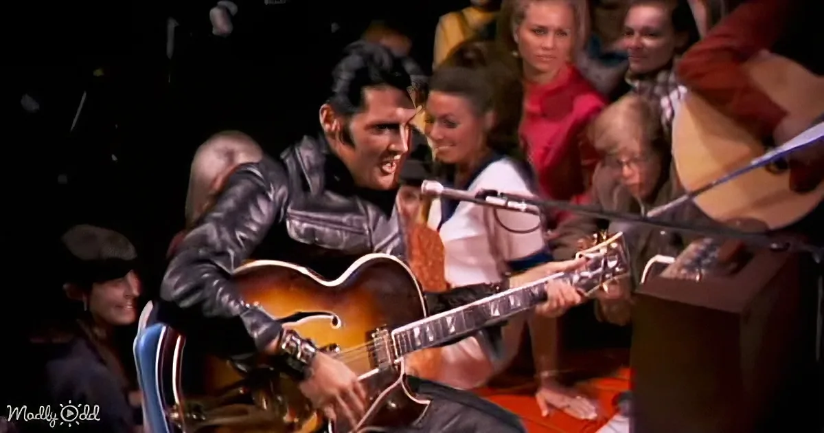 Elvis Presley in a leather jacket plays guitar on stage, surrounded by a crowd.