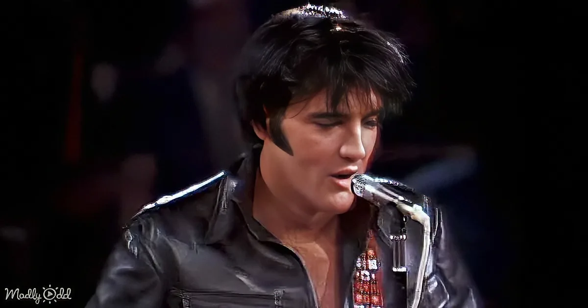 A person in a black leather outfit sings into a microphone, wearing a headband, with a dark background.