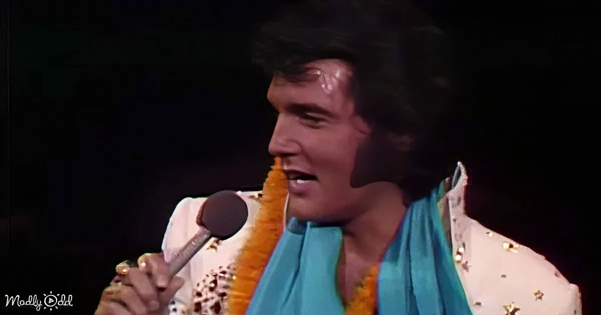 Elvis Presley in a white sequined suit with a blue scarf, holding a microphone, singing on stage.