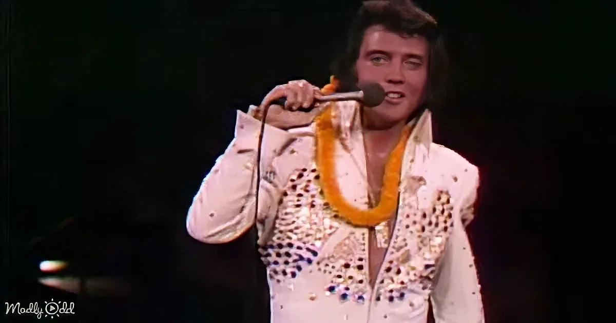 A man in a white sequined suit with a microphone and a lei around his neck is performing on stage.