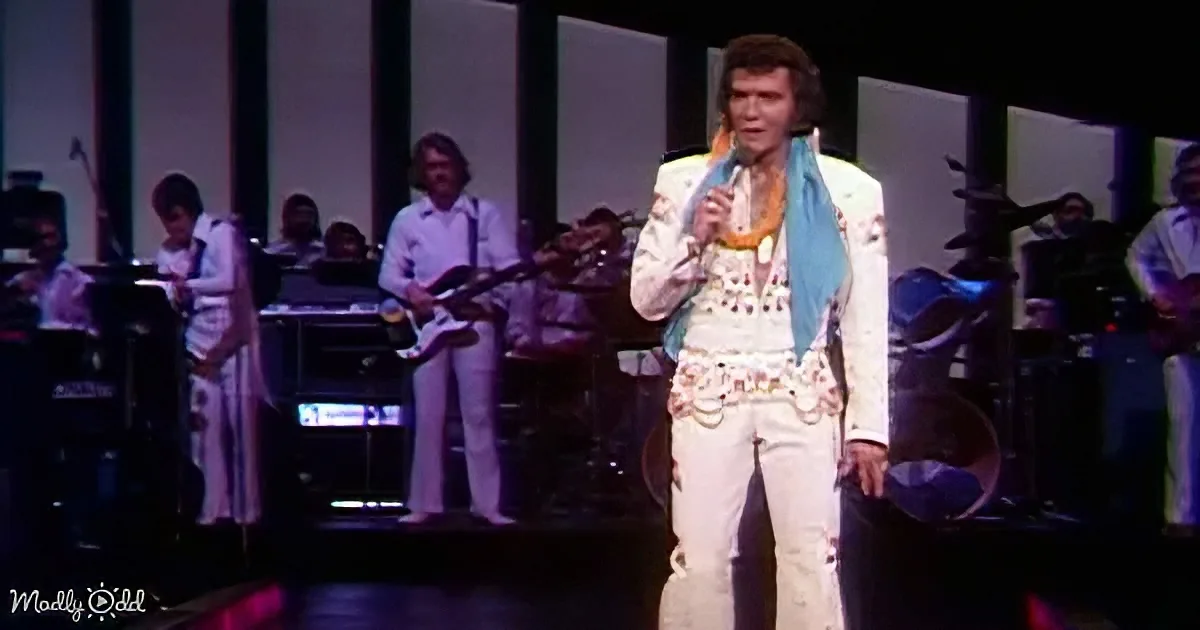 A performer in a white outfit with a blue scarf sings on stage, accompanied by a band.