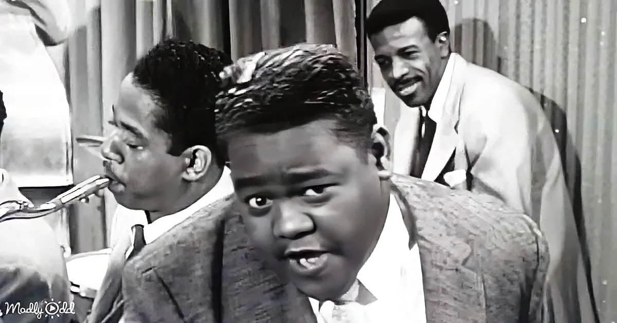 A man in a suit and tie is singing, with a saxophonist and another man in the background.