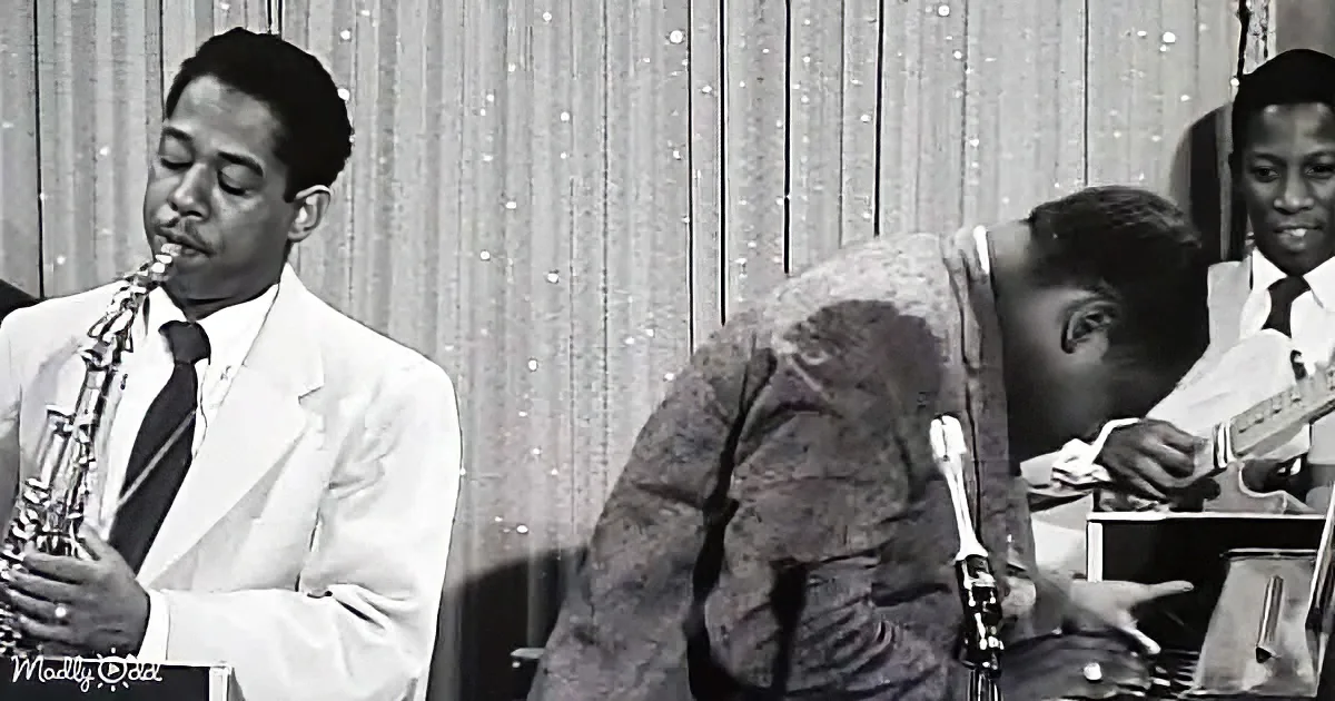 A man in a white suit plays saxophone while another bends over, possibly adjusting something.