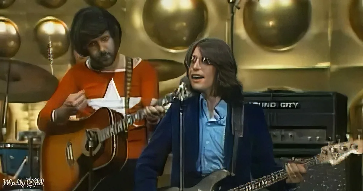 A man in a blue jacket plays guitar while another in an orange shirt plays acoustic guitar in a music studio.