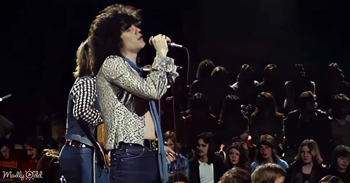 A man with long hair sings into a microphone on stage, surrounded by a crowd and musicians.
