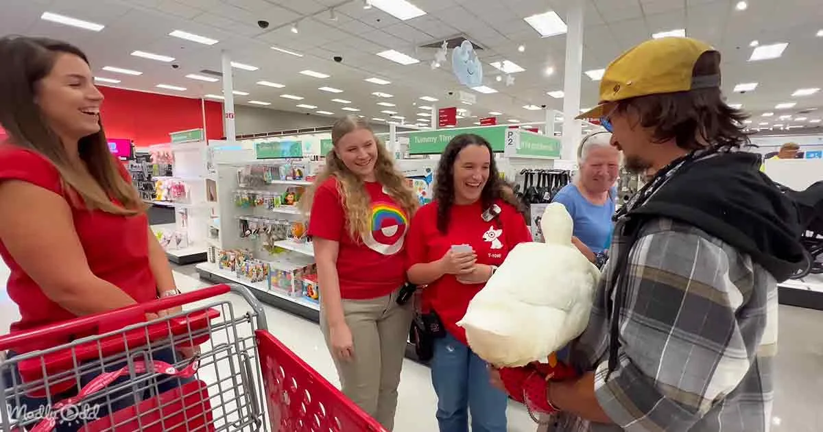 A man in a plaid shirt holds a large white stuffed animal, interacting with three women in red shirts in a Target store.