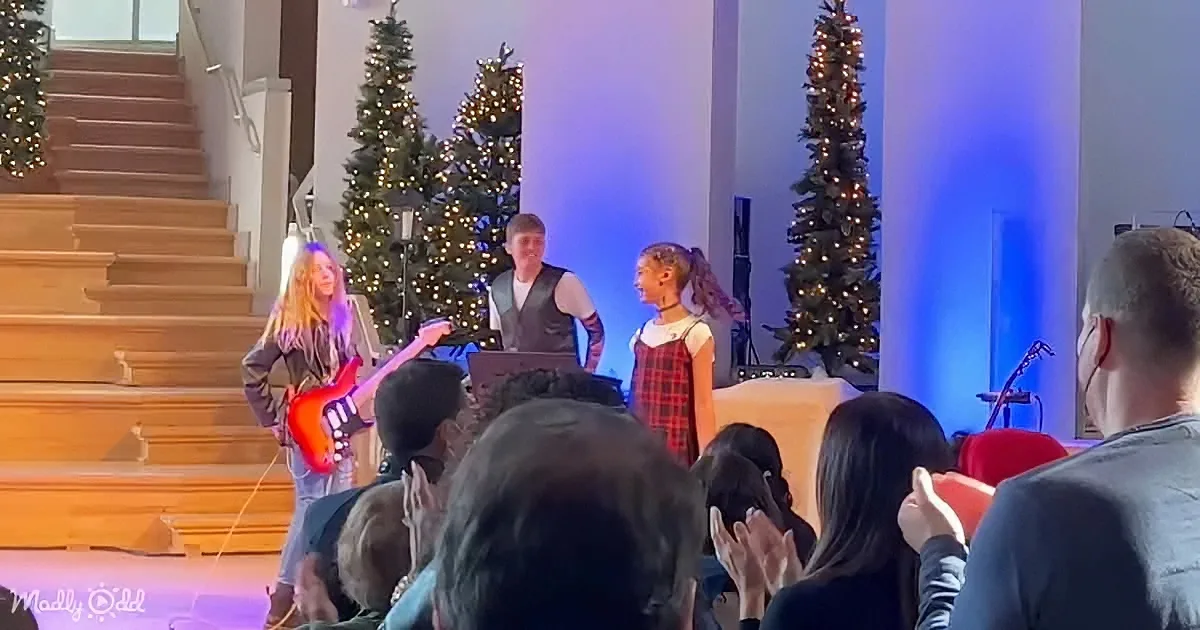 A young girl plays guitar on stage with others, surrounded by Christmas trees and an audience clapping.