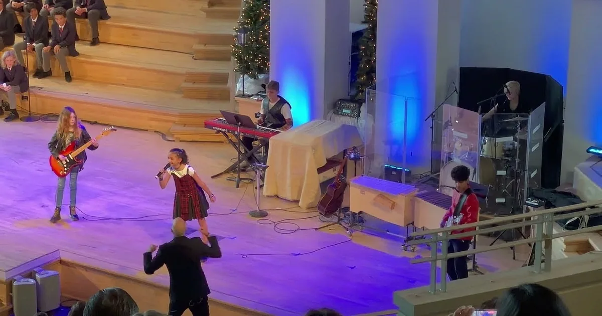 A young girl sings into a microphone on stage, accompanied by a guitarist and keyboardist.