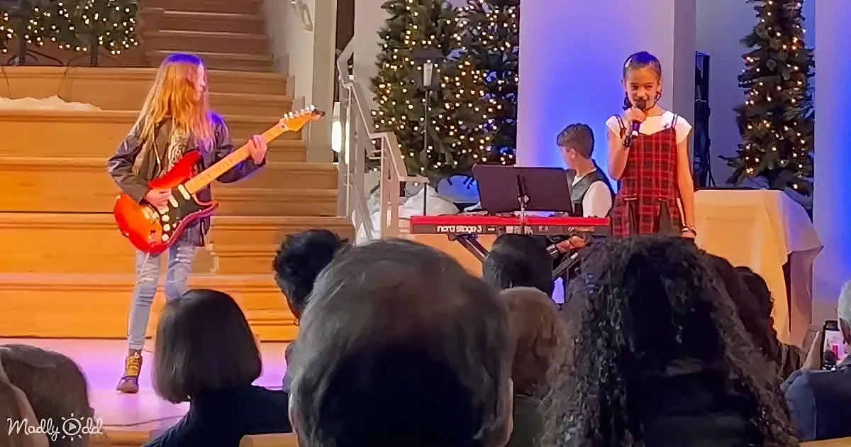 A young girl sings into a microphone while a boy plays guitar on stage, surrounded by Christmas trees and an audience.
