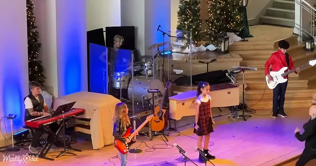 A young girl sings on stage with a band, surrounded by Christmas trees and colorful lighting.
