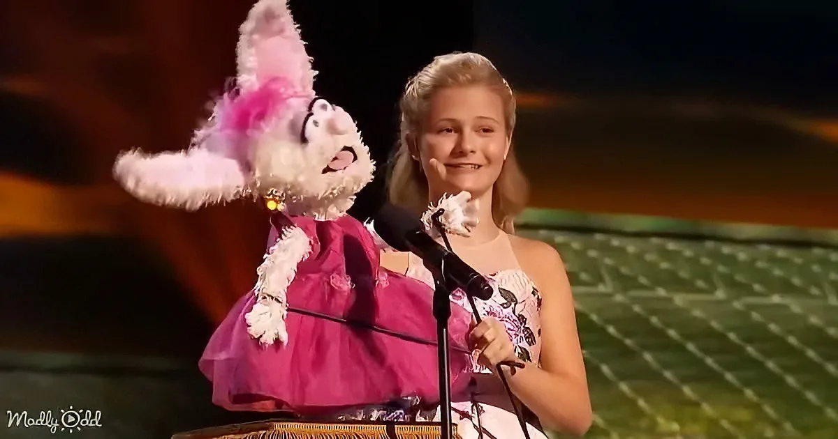 A young girl in a floral dress holds a microphone and puppet on stage.