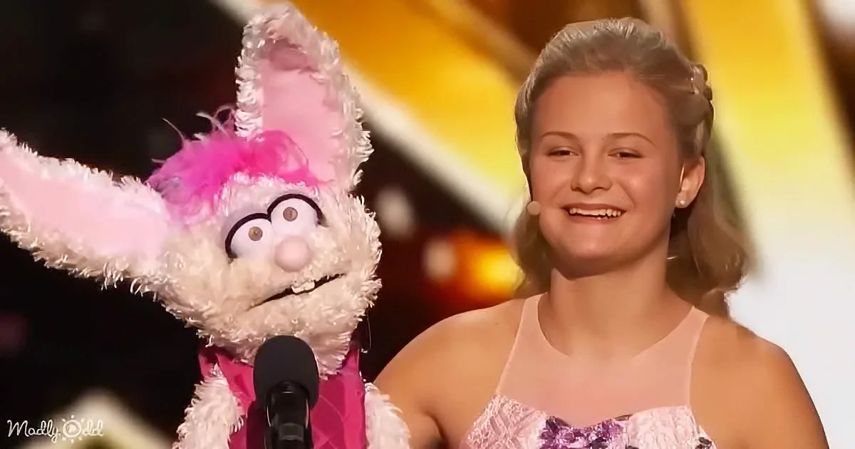 A young girl in a pink dress smiles next to a puppet with pink ears and a microphone.