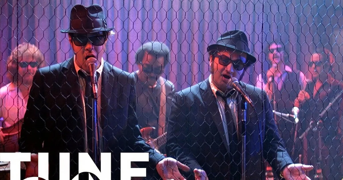Two men in suits and hats sing into microphones, surrounded by a band and a chicken wire backdrop.