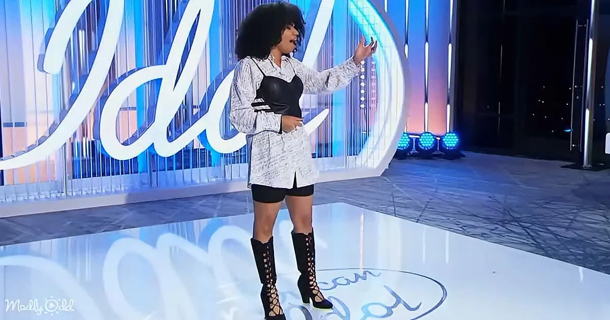 A person with an afro, wearing a white and black outfit, stands on a stage with a "Idol" backdrop, gesturing with one...