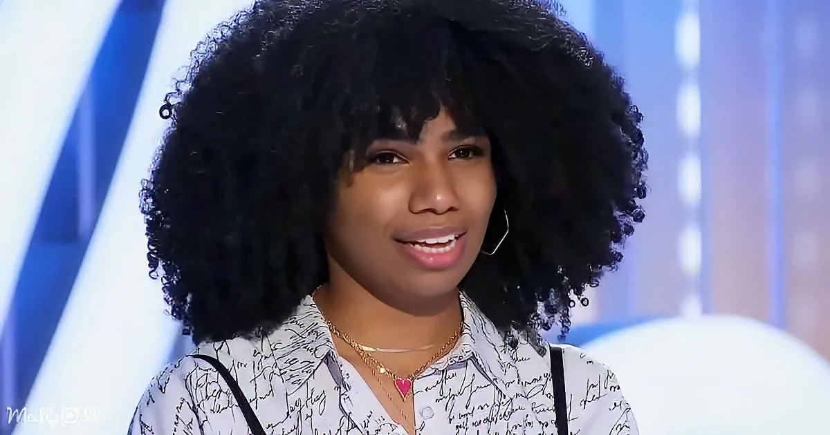 A person with a large, curly afro hairstyle, wearing a white shirt with black patterns and a gold necklace, smiles while...