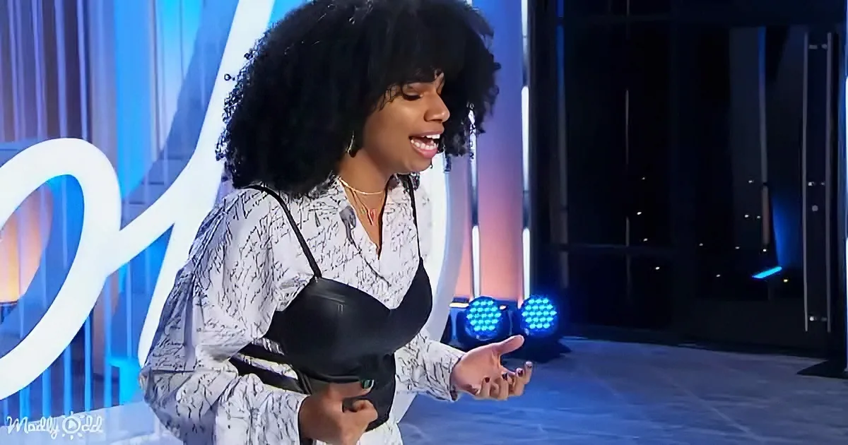 A woman with curly hair, wearing a white patterned shirt and black bra, stands on a stage with blue lighting.