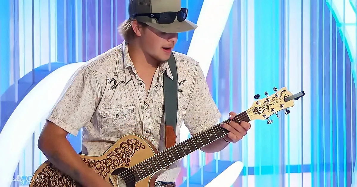A man in a cowboy hat and floral shirt plays a guitar on stage with a colorful background.