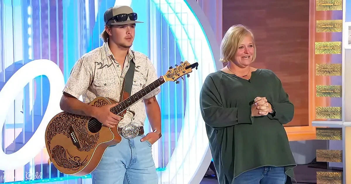 A man in a hat and shirt holds a guitar, standing next to a woman in a green top on a brightly lit stage.
