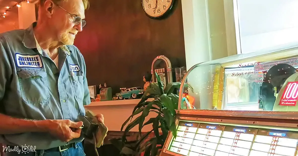 An older man in a "Jukeboxes Unlimited" shirt stands near a vintage jukebox, holding a cloth, with a plant and clock in...