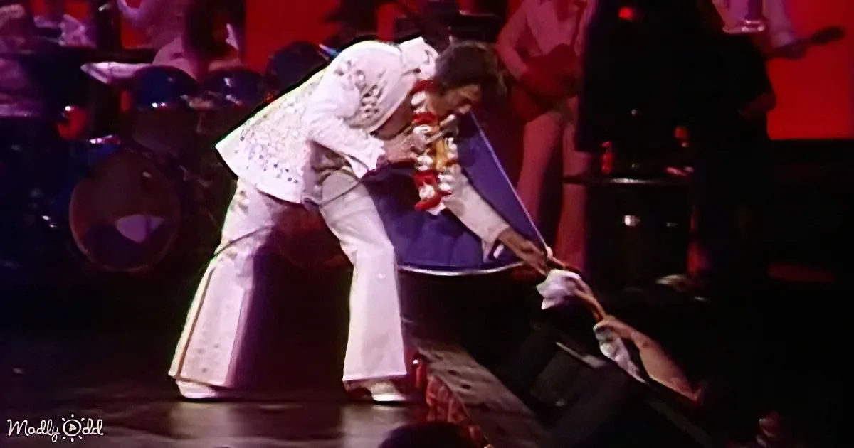 A performer in a white suit is bending down, holding a microphone, with a blue cape draped over their shoulder.